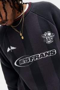 Svart texturerad tröja med röd randig halsringning, med en vit logotyp och text, samt lager av guldkedjor med hängen.