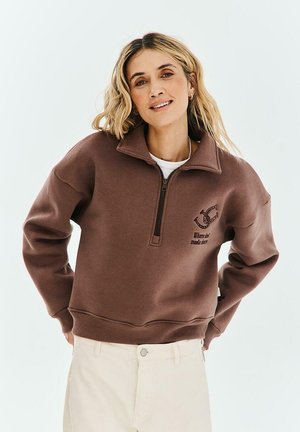 Uśmiechnięta kobieta o blond włosach nosi brązową bluzę z zamkiem do ćwiartki i białe spodnie, pozuje z rękami za plecami.