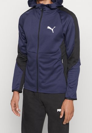 Mand iført navy og sort Puma hættetrøje med lynlås med hvidt logo på brystet og sorte Puma joggingbukser med hvidt logo på låret.