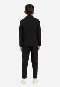 Traje negro con una chaqueta entallada y pantalones rectos, que presenta una textura suave y un brillo sutil, combinados con zapatillas blancas.