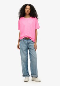 Camiseta de manga corta rosa brillante con un ajuste relajado, combinada con unos pantalones vaqueros rectos de color azul claro y zapatillas blancas. Detalle de cinturón sencillo.