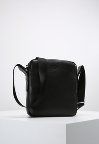 Borsa a tracolla nera in pelle, di forma quadrata, superficie testurizzata, tracolla regolabile e un logo sobrio impresso sul davanti.