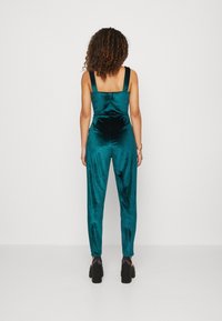 Teal sammet jumpsuit med breda axelremmar, figursydd överdel och avsmalnande ben. Har en synlig bakseam och svarta klackskor med blockklack.