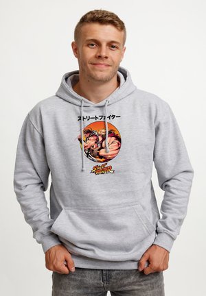 Mand iført lysegrå hættetrøje med Street Fighter-grafik af to figurer, der kæmper inde i en orange cirkel, og tekst over og under.