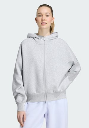 Ung kvinde iført lysegrå lynlåshættetrøje med hætte og hvide bukser, stående med den ene hånd på hoften mod en ensfarvet baggrund.