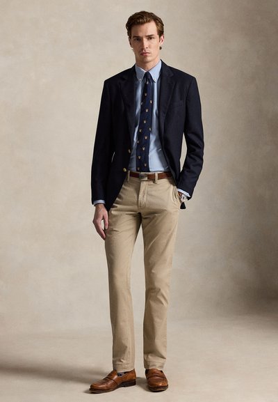 Bărbat purtând sacou bleumarin, cămașă albastră cu nasturi, cravată bleumarin cu model, pantaloni bej, curea maro și mocasini maro din piele, stând cu mâna în buzunar.
