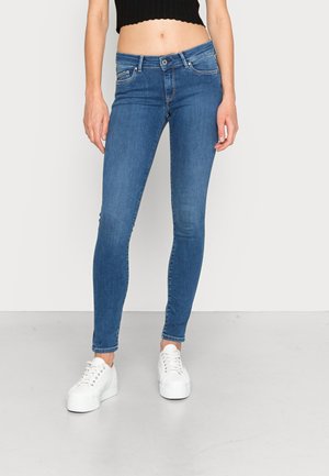 Frau trägt blaue Skinny-Jeans mit mittlerer Taille und weiße Plateau-Sneakers, steht vor einem schlichten hellen Hintergrund.