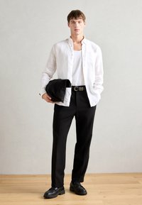 Camisa de linho branca de botões sobre uma t-shirt branca, calças pretas de cintura alta, cinto preto com fivela quadrada, segurando uma clutch preta, sapatos escuros.