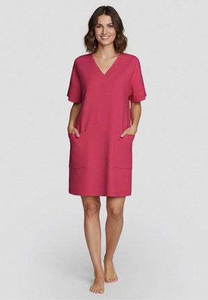 Nainen seisoo paljain jaloin, yllään lyhythihainen, polvipituinen vaaleanpunainen mekko, jossa V-kaula-aukko ja etutaskut, hymyillen yksinkertaista taustaa vasten.