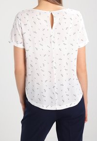 Let, hvid bluse med et diskret mønster af små sorte accenter. Har korte ærmer, en rundet nederkant og en nøglehuldsdetalje bagpå.