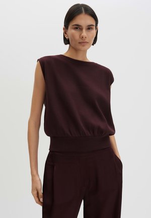 Donna con capelli corti e scuri indossa un top senza maniche bordeaux scuro e pantaloni abbinati, in piedi davanti a uno sfondo chiaro uniforme.