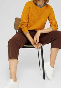 Personne assise sur une chaise portant un pull orange texturé, un pantalon marron raccourci et des baskets blanches sur un fond uni.