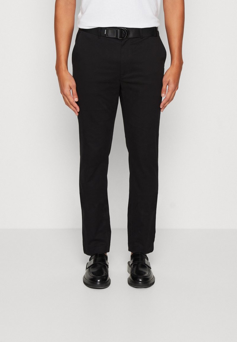 Schwarze Hosen mit schmaler Passform aus glattem Stoff, versehen mit einem Gürtel mit rundem Schnallenverschluss, kombiniert mit schwarzen Schuhen. Einfaches Design, ohne Muster.