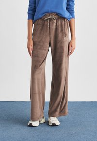 Άτομο που φορά καφέ παντελόνι βελουτέ φαρδιάς γραμμής με κορδόνι στη μέση και λευκά αθλητικά παπούτσια, στέκεται σε μπλε χαλί.