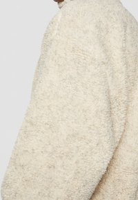Giacca in tessuto peloso color crema, con superficie testurizzata e soffice, vestibilità ampia, collo rotondo e spalle senza cuciture.