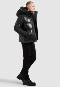 Chaqueta acolchada negra con una gran capucha, material brillante, cierre de cremallera y dos bolsillos laterales; combinada con pantalones negros y botas negras robustas.