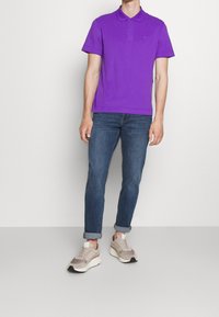 Camisa polo de algodón morada con cuello y dos botones, combinada con jeans de mezclilla azul remangados y zapatillas neutras. Estilo simple y casual.