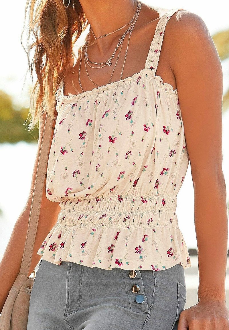 Kobieta w kwiecistej, bezrękawnikowej bluzce z marszczonym pasem i warstwowych srebrnych naszyjnikach, niosąca beżową torbę na ramię, na zewnątrz w świetle dziennym.