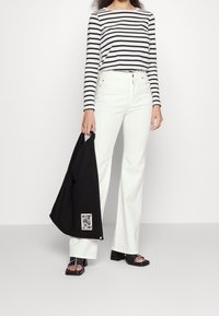 Gestreiftes langärmliges Hemd in Weiß und Marineblau, kombiniert mit weißen weit geschnittenen Jeans. Hält eine schwarze Tragetasche mit grafischem Detail. Schwarze hohe Sandalen.