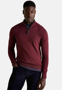 Bordový sveter so zipsom a rebrovanými manžetami a spodným lemom, vyrobený z mäkkej tkaniny; navrstvený na modrej pruhovanej košeli. Na hrudi má logo.