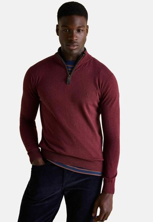 Weinrot zip-neck Pullover mit gerippten Bündchen und Saum, aus weichem Material; über einem blau gestreiften Hemd getragen. Mit einem Logo auf der Brust.