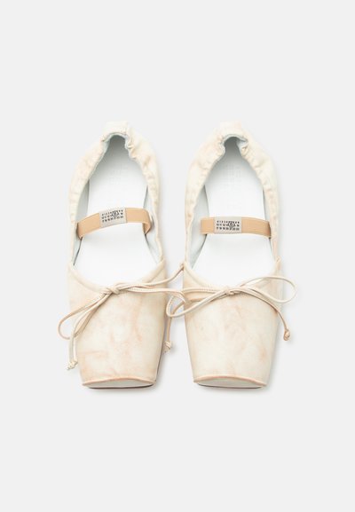 Par bež baletnih copatov s kvadratnimi konicami, elastičnimi trakovi okoli gležnjev in pentljami, prikazanih od zgoraj na belem ozadju.