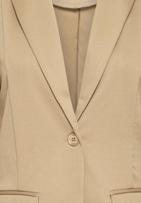 Blazer beige con revers a punta, chiusura a singolo bottone, tessuto texturizzato e tasca frontale sul lato sinistro. Fodera interna liscia.