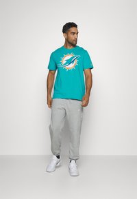 Türkisfarbenes T-Shirt mit einem Miami Dolphins Logo, kombiniert mit hellgrauen Jogginghosen und weißen Sportschuhen. Einfaches Rundhals-Design.