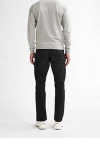 Hombre de pie de espaldas, vistiendo una sudadera gris, jeans negros y zapatillas blancas, sobre un fondo blanco liso.