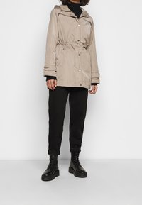 Béžová parka s pružným pasem, zipem a druky, a kapucí. Nositá přes černé kalhoty a černé šněrovací boty.