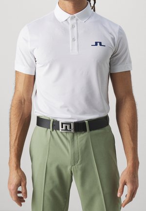 Männlicher Torso trägt ein weißes kurzärmeliges Poloshirt mit blauem Logo, hellgrüne Hose und einen schwarzen Gürtel mit silberner geometrischer Schnalle.