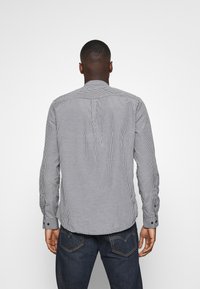 Camisa de cuadros en blanco y negro con cuello abotonado y mangas largas, que presenta un pliegue en la parte posterior y botones negros en los puños.