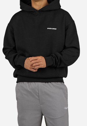 Person trägt einen schwarzen Kapuzenpullover mit "PEGADOR"-Logo und graue Hose mit kleinem weißem Schriftzug am linken Oberschenkel.