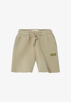 Dijete odjeveno u bež kratke hlače s vezicama s logotipom "American Vintage", maslinasto zelenu majicu dugih rukava i plave cipele bez vezica stoji na svijetloj podlozi.