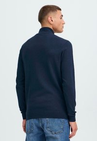 Pulover din tricot de culoare bleu marin, cu guler înalt, manșete și tiv cu dantelă, purtat peste blugi de denim de culoare albastru deschis. Are un design simplu.