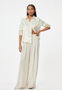 Camisa de seda crema de botones con mangas remangadas, combinada con pantalones de talle alto y pierna ancha con pliegues en un color crema a juego.