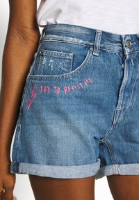 Džínové šortky s opotřebovaným vzhledem, světle modré barvy, s manžetou na spodním okraji a růžovým vyšívaným textem "TRY TO RESIST ME."