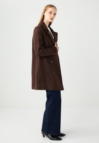 Brauner, doppelt geknöpfter Mantel mit Reverskragen, aus dickem Stoff. Kombiniert mit dunkelblauen, ausgestellten Jeans und schwarzen, spitzen High Heels.