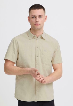 Ung mand med kort hår iført en beige kortærmet skjorte, stående mod en ensfarvet lys baggrund.