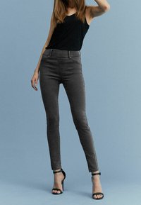 Frau in schwarzem ärmellosem Top, eng anliegenden dunkelgrauen Jeans und schwarzen High-Heels, die vor einem schlichten blauen Hintergrund steht.