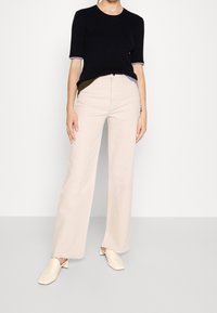 Beige Hosen mit weitem Bein kombiniert mit einem enganliegenden schwarzen Pullover mit kontrastierenden, gerippten Bündchen. Die Schuhe sind cremefarben mit einem Blockabsatz.