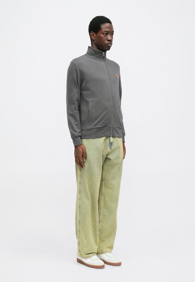 Gray zip-up jacket with high collar, two side pockets, and small orange emblem; paired with light green wide-leg pants and white shoes.