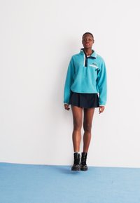 Tyrkysový fleece pulóver s čiernym golierom a bielou náprsné vreckom, spárovaný s čiernou sukňou a čiernymi bojovými topánkami, na bielom pozadí.