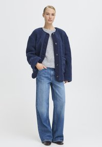 Veste en polaire bleu marine avec une surface texturée, col arrondi et grands boutons, associée à un jean large bleu clair et un pull gris.