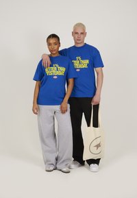 Tricouri din bumbac albastre cu text galben "1% MA BINE DECÂT IERI", pantaloni de trening gri și o geantă de toată ziua bej cu logo, purtate de două persoane.