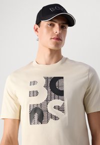 Young man wearing a black cap and a beige t-shirt with a large black and patterned abstract design on the front.