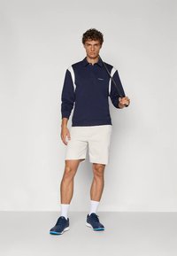 Tmavomodré polokošeľa s dlhým rukávom s bielymi detailmi, krémové nohavice a tmavomodré topánky. Hladká textúra, priliehavý dizajn a golier. Golfová palica v ruke.