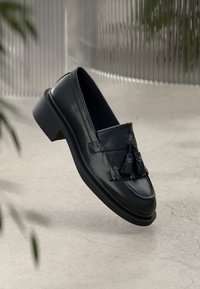 Svart skinnloafers med frynser, glatt tekstur, rund tå og grov hæl. Minimalistisk design mot en nøytral bakgrunn.