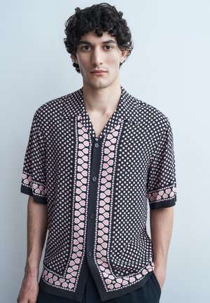 Νέος άνδρας με σγουρά σκούρα μαλλιά, ντυμένος με μαύρο κοντομάνικο πουκάμισο με μοτίβο από μικρές και μεγάλες ρόζ καρδιές, στέκεται μπροστά σε απλό φωτεινό φόντο.