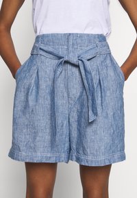 Světle modré džínové kraťasy s vysokým pasem na pásek, bočními kapsami a texturovanou tkaninou. Mají ohrnutý lem a kontrastní steh.